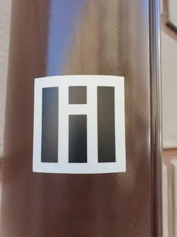 A square sticker with a simple design featuring three black vertical bars of varying heights, set against a white background. It is affixed to a brown cylindrical surface, possibly a pole or pillar.