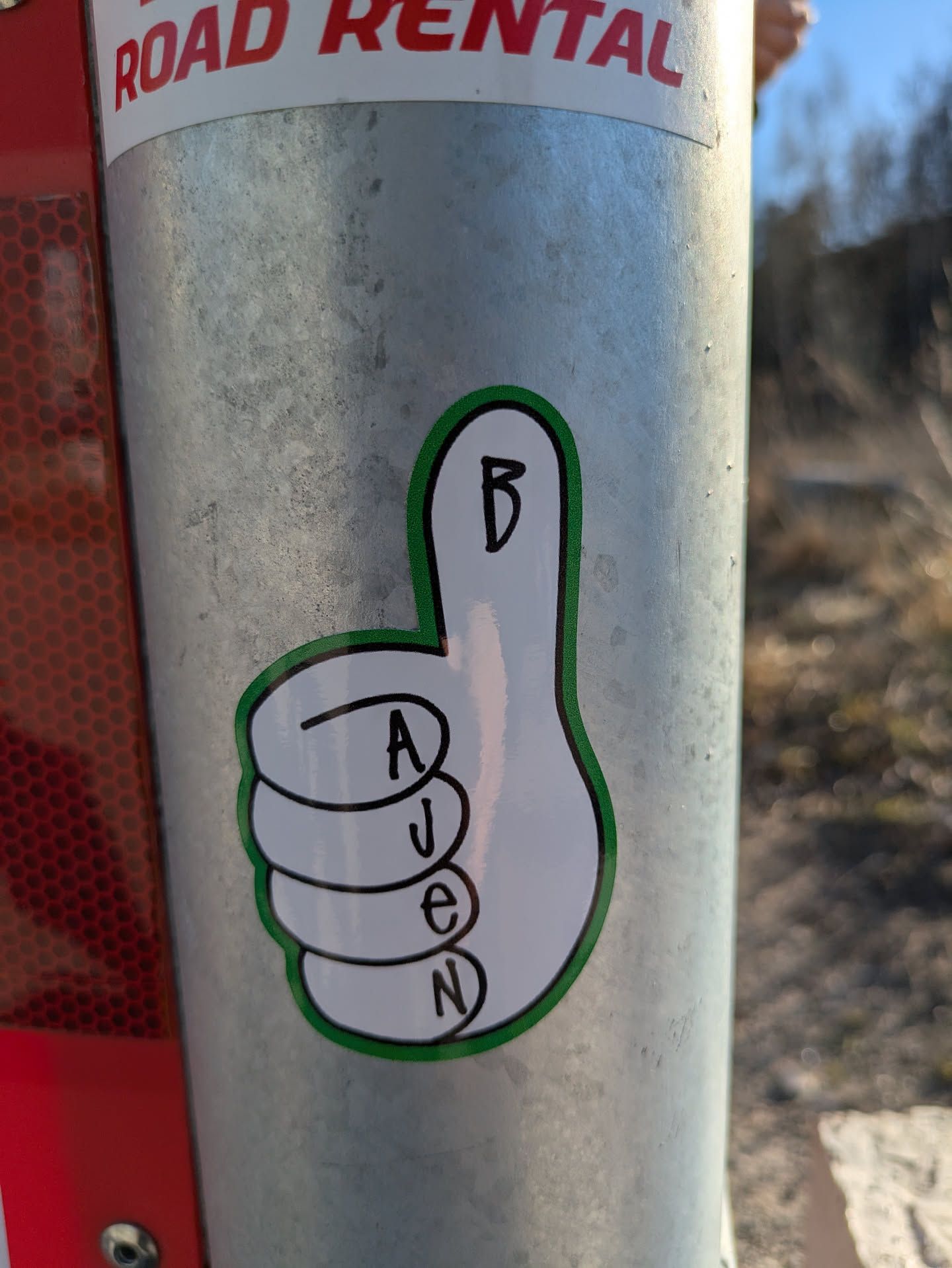 A thumbs-up sticker with a green outline and the word "AJENB" spelled vertically.  The sticker is adhered to a metal post.
