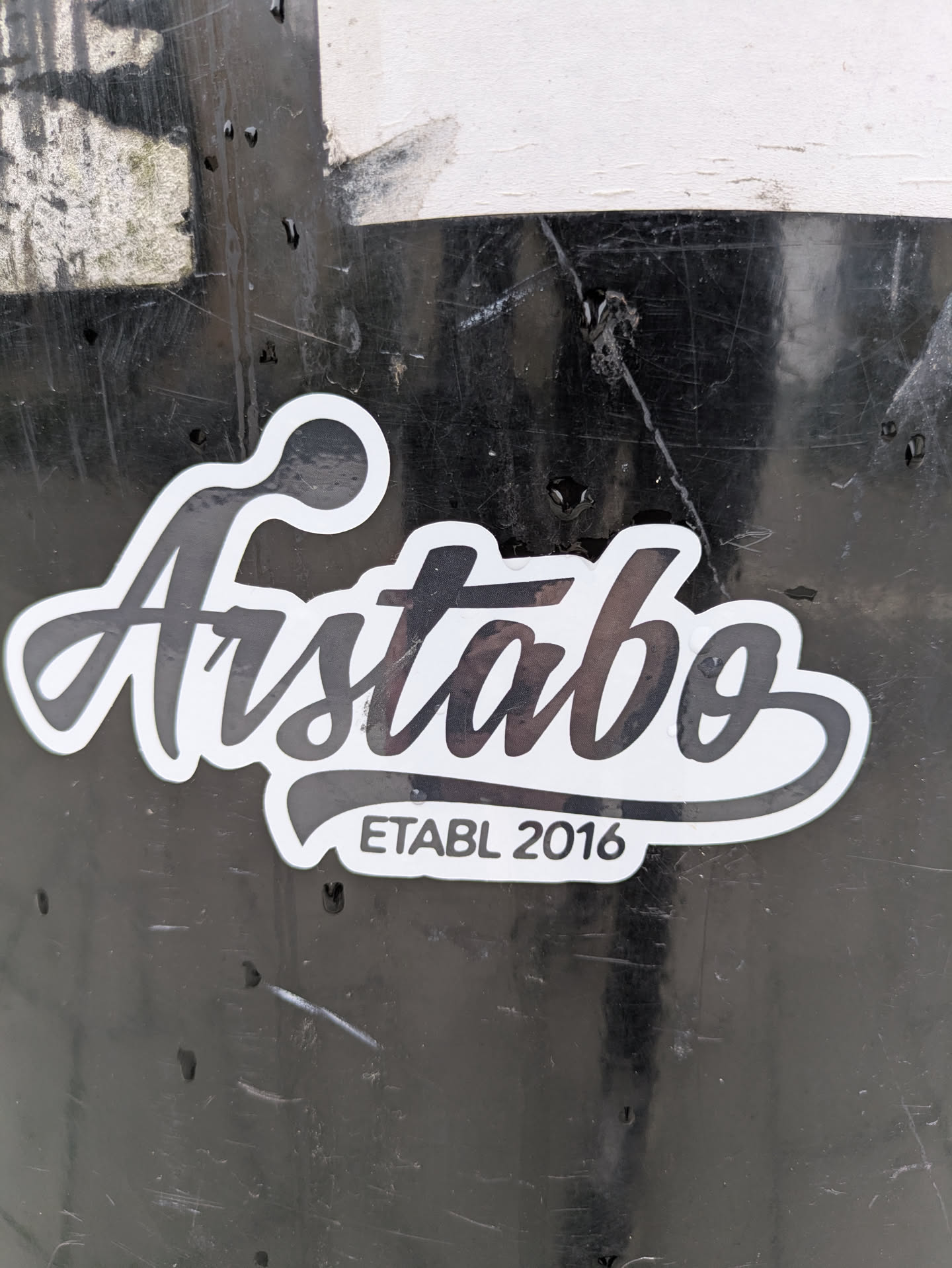 The sticker features a prominent, stylized word "Aistabo" in a flowing, elegant script font. It is primarily black with a clean white outline, creating a strong contrast against the dark, scratched background it's affixed to. Below the main text, in a smaller, sans-serif font, are the words "ETABL 2016" on a small, curved banner-like shape, also in black text on a white background with a black outline. The overall shape of the sticker follows the contours of the text.