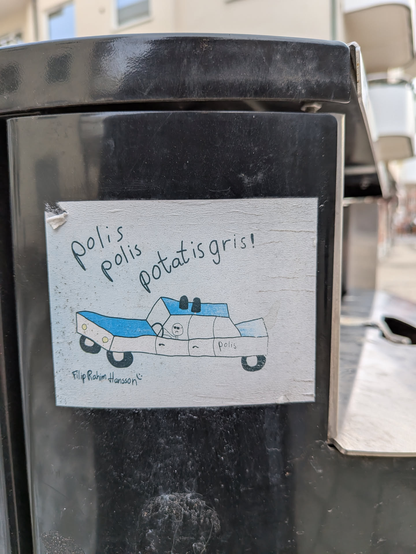 A rectangular, hand-drawn sticker featuring a cartoonish police car on a white background. The car is outlined in black, with light blue details on the roof lights and front section, and yellow headlights. A small, simple figure with a sad expression is visible in the driver's seat. Above the car, the text 'Polis polis potatis gris!' is written in black, and below, 'Filip Rahim Hansson' with a small smiley face. The sticker shows signs of wear, particularly a slight tear on the top-left corner, and is affixed to a dark, textured surface.