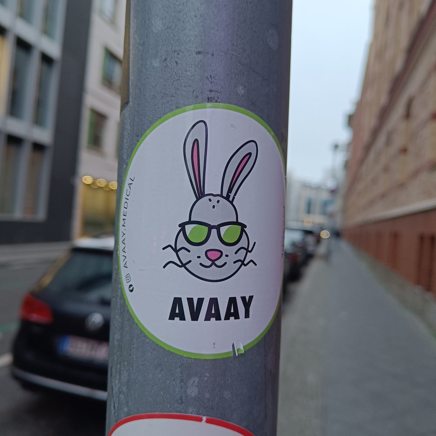 A round sticker featuring a cartoon rabbit wearing sunglasses. The text 'AVAAAY' is written below the rabbit.  The sticker is attached to a metal pole on a city street.