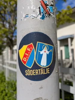 This round sticker features two shields. The left shield has the letters "D.I.F." in white on a blue diagonal stripe over a yellow background. The right shield depicts a robed figure with a halo holding a staff, set against a blue background. The word "SÖDERTÄLJE" is printed below the shields.