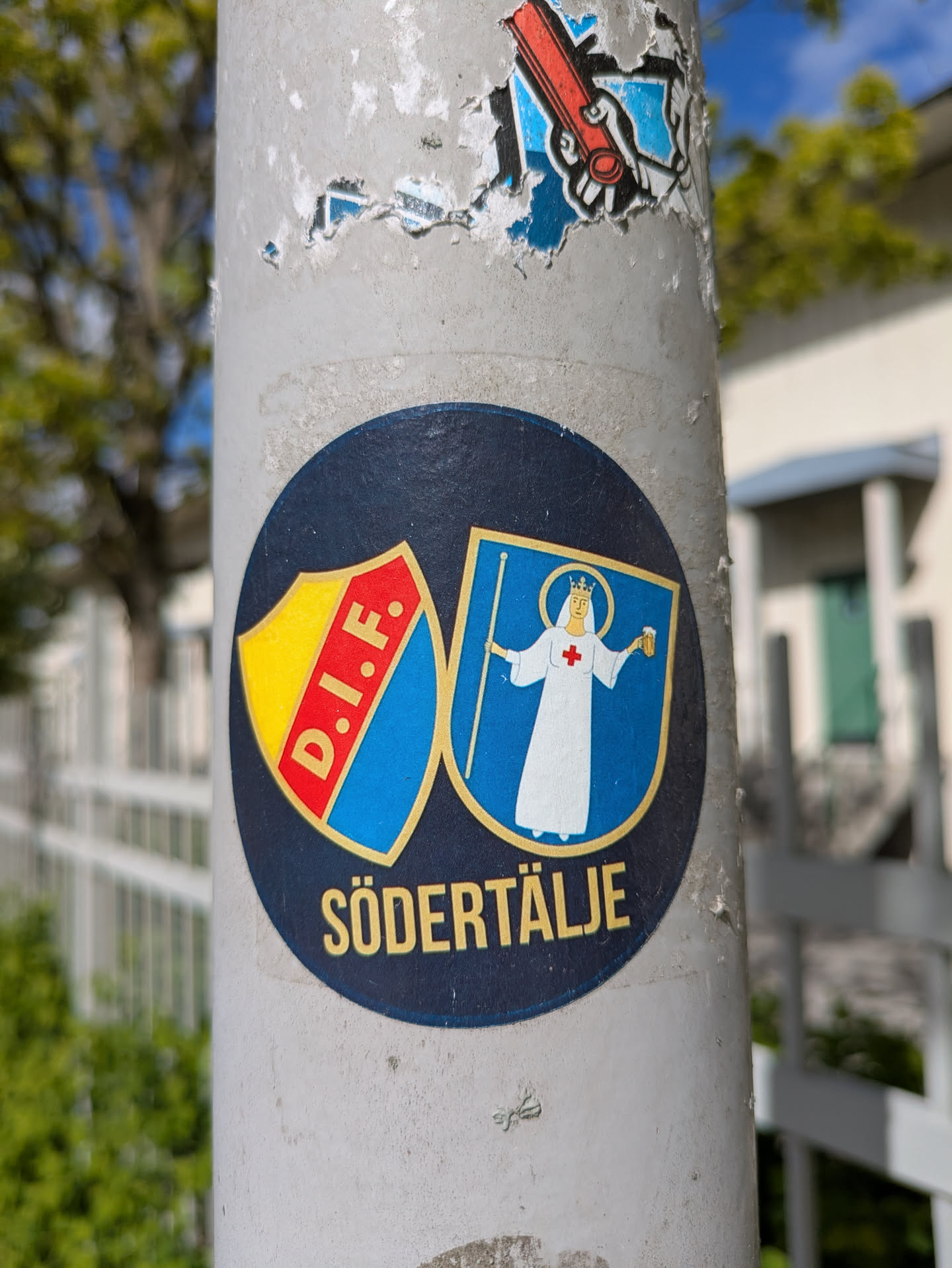 This round sticker features two shields. The left shield has the letters "D.I.F." in white on a blue diagonal stripe over a yellow background. The right shield depicts a robed figure with a halo holding a staff, set against a blue background. The word "SÖDERTÄLJE" is printed below the shields.