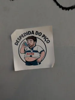 A sticker depicts a cartoon man with a slightly overweight build, wearing a sports shirt with 'KTM' emblazoned on it. He's using an inhaler and encircled by the text 'DESPEDIDA DO PIÇO'.