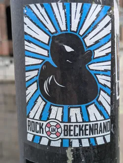 A sticker featuring a stylized rubber duck in black and blue color scheme. The duck is depicted in the center, surrounded by a sunburst pattern. The words "Rock am Beckenrand" are printed at the bottom. 