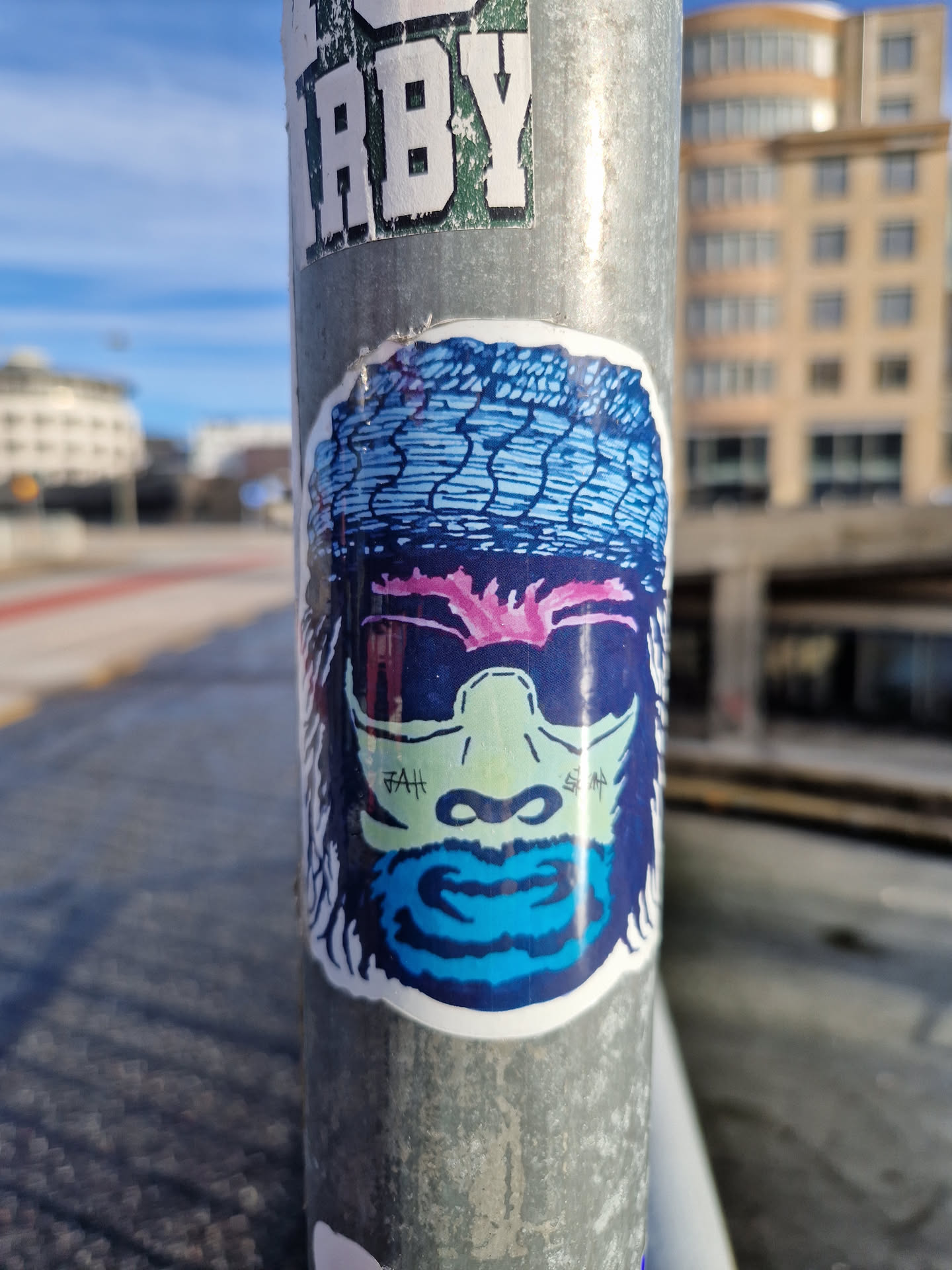 A sticker featuring a stylized illustration of a monkey's face. The monkey is wearing a bandana and sunglasses. The style is bold and graphic with vibrant colors and a slightly psychedelic feel. The sticker is affixed to a metal pole, showing some wear from the elements.