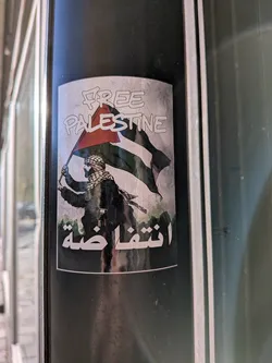 The sticker features a stylized illustration of a person wearing a keffiyeh and holding the Palestinian flag aloft. The flag's colors (black, white, green, and red) are prominent. Below the figure, in Arabic script, is the word 'Intifada'. The overall aesthetic is somewhat gritty, with a textured background that gives it a worn or distressed look. The sticker is oval-shaped with a white border.