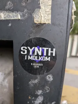 A circular sticker with a black background. The prominent text 'SYNTH' is in large, white, sans-serif capital letters, with the 'TH' partially overlaying a stylized purple gear or cogwheel. Below this, in smaller white text, reads 'I MOLKOM'. At the bottom of the sticker, the date '9 AUGUSTI 2025' is also written in white text.