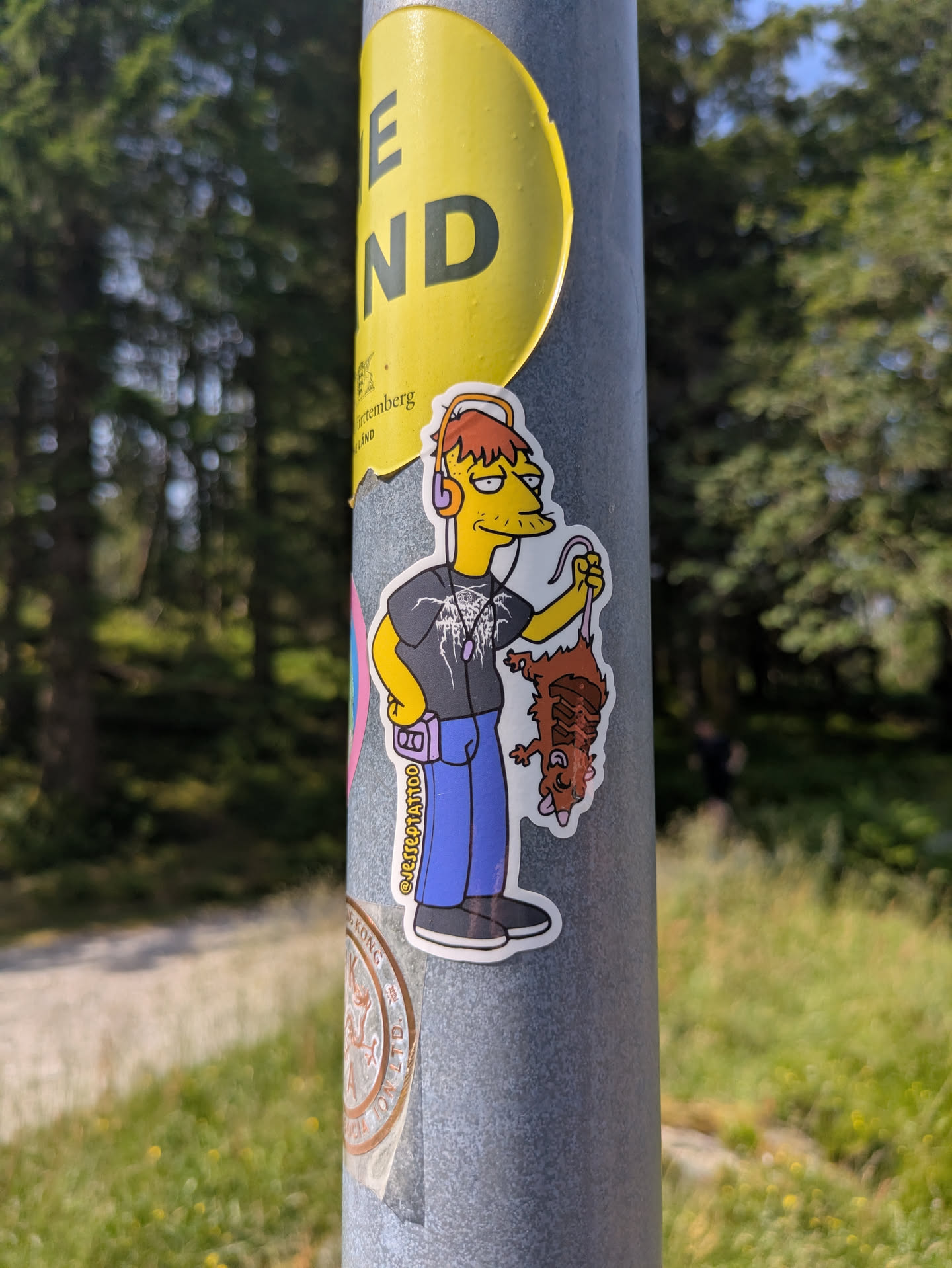 A white vinyl sticker affixed to a grey metal pole features a yellow-skinned cartoon character with a large, pointed nose and mustache, and spiky orange hair. He wears yellow over-ear headphones, a dark t-shirt with a detailed white graphic resembling a heavy metal band logo, blue pants, and black sneakers. A purple portable cassette player (Walkman) is clipped to his belt, and purple earbuds hang from his neck. The character is shown holding a dead, brown, and partially eviscerated rodent (possibly a rat or possum) by its tail, which is hooked. The background is blurred, showing green trees and foliage under a bright sky.