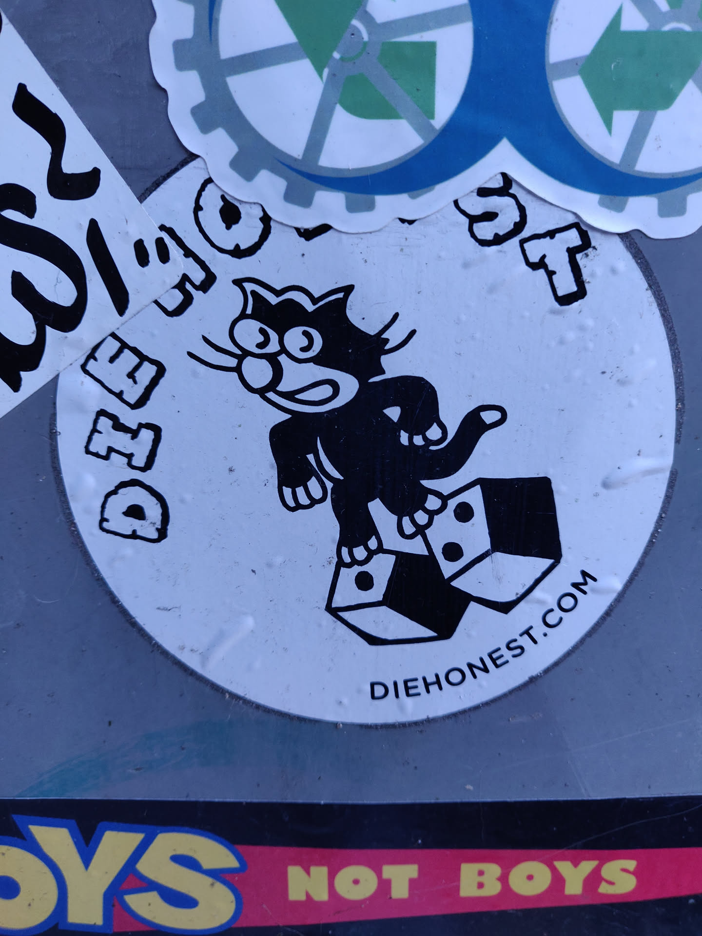 Round sticker featuring a cartoon cat standing on top of two dice. The text "DIE HONEST" is visible around the image, along with the website "DIEHONEST.COM" at the bottom. The style is reminiscent of vintage cartoons or graphic design.
