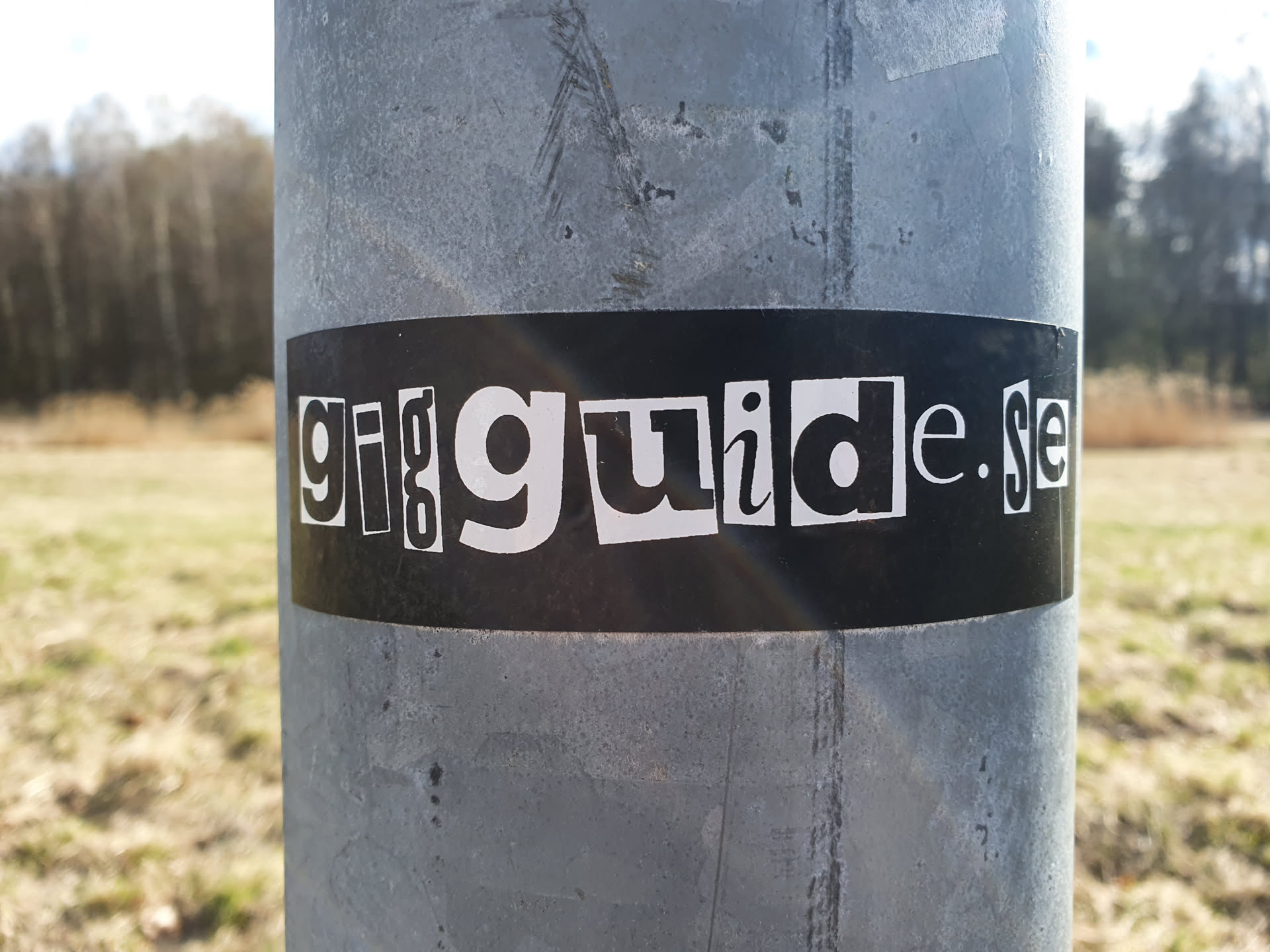 A black and white sticker with the text "gigguide.se" on a black background. The letters are arranged in a slightly irregular way, giving it a collage-like appearance. The sticker is affixed to a gray metal pole outdoors, with a blurred background of a field and trees.