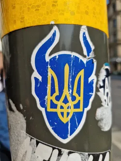 This image shows a sticker affixed to a pole. The sticker is weathered and damaged, showing signs of wear and tear. It displays the Ukrainian Tryzub, a prominent national symbol. The design incorporates horns around the Tryzub giving it a more aggressive, defiant look. The colors are blue and gold, representing the Ukrainian flag. The background is a greyish-black surface.