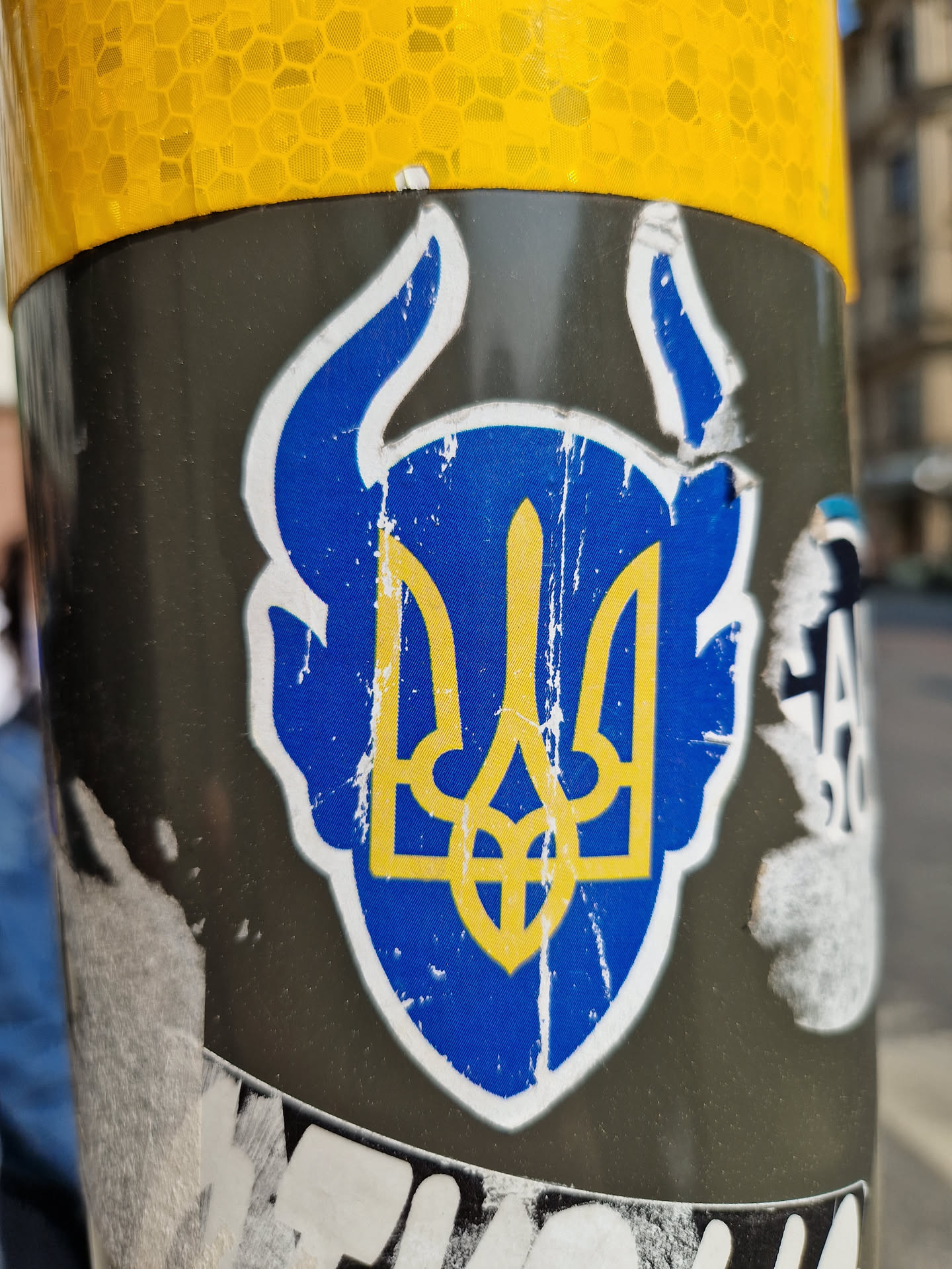 This image shows a sticker affixed to a pole. The sticker is weathered and damaged, showing signs of wear and tear. It displays the Ukrainian Tryzub, a prominent national symbol. The design incorporates horns around the Tryzub giving it a more aggressive, defiant look. The colors are blue and gold, representing the Ukrainian flag. The background is a greyish-black surface.