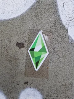 A green diamond-shaped sticker featuring the iconic Sims logo. It's affixed to a textured gray surface. 
