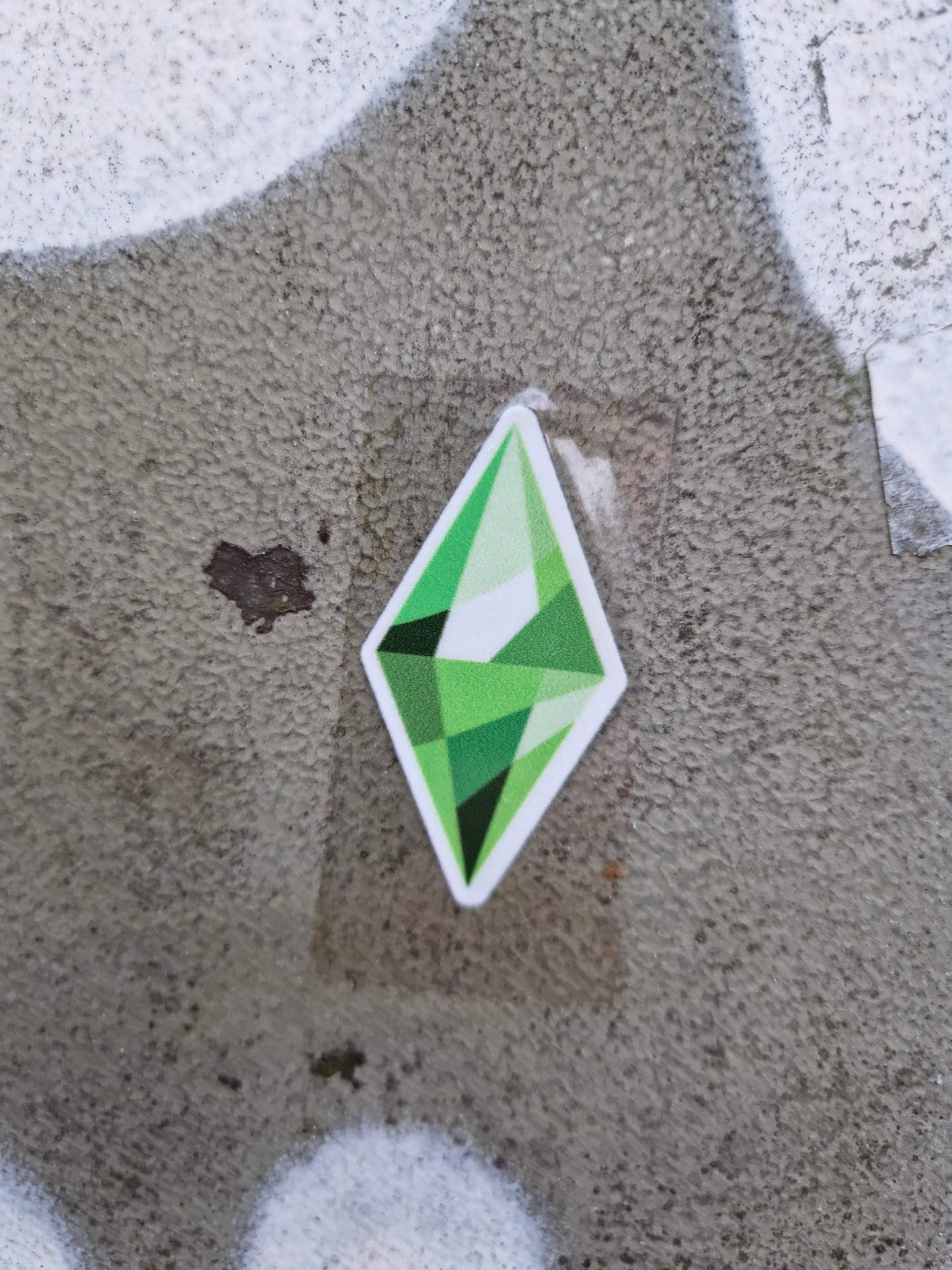 A green diamond-shaped sticker featuring the iconic Sims logo. It's affixed to a textured gray surface. 