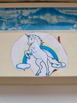 This sticker features a charming illustration of a light blue unicorn elegantly standing on its hind legs, set against a vibrant rainbow backdrop. Soft white clouds are depicted beneath the unicorn, adding to the whimsical scene. The style is simple yet captivating, conveying a sense of magic and fantasy.