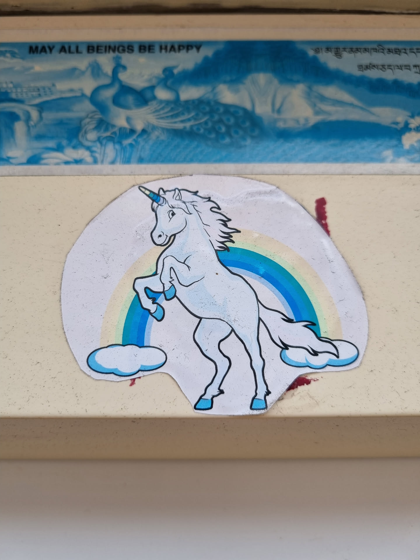 This sticker features a charming illustration of a light blue unicorn elegantly standing on its hind legs, set against a vibrant rainbow backdrop. Soft white clouds are depicted beneath the unicorn, adding to the whimsical scene. The style is simple yet captivating, conveying a sense of magic and fantasy.