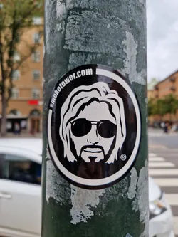 Black and white sticker featuring a stylized portrait of a man with long hair and sunglasses. The text "smoketower.com" is visible at the top of the sticker. The sticker is affixed to a weathered green pole.