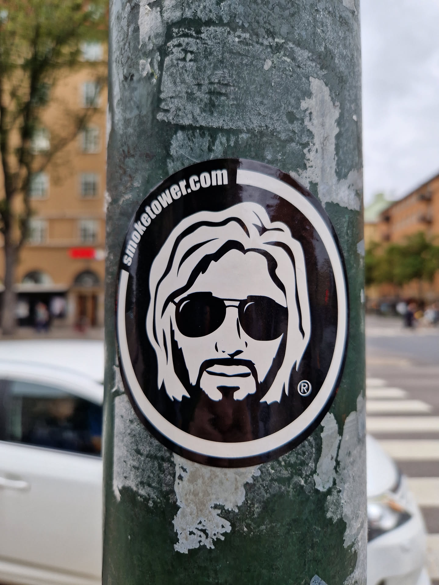 Black and white sticker featuring a stylized portrait of a man with long hair and sunglasses. The text "smoketower.com" is visible at the top of the sticker. The sticker is affixed to a weathered green pole.