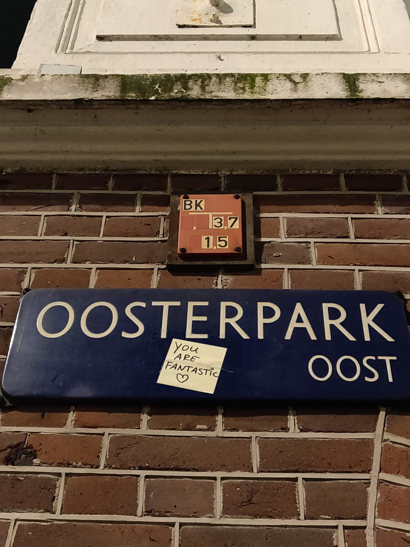A photograph of a blue street sign that reads "Oosterpark Oost." Affixed to the sign is a small yellow sticky note with the handwritten message: "You are fantastic." There is also a small orange metal box above the street sign with some numbers on it.