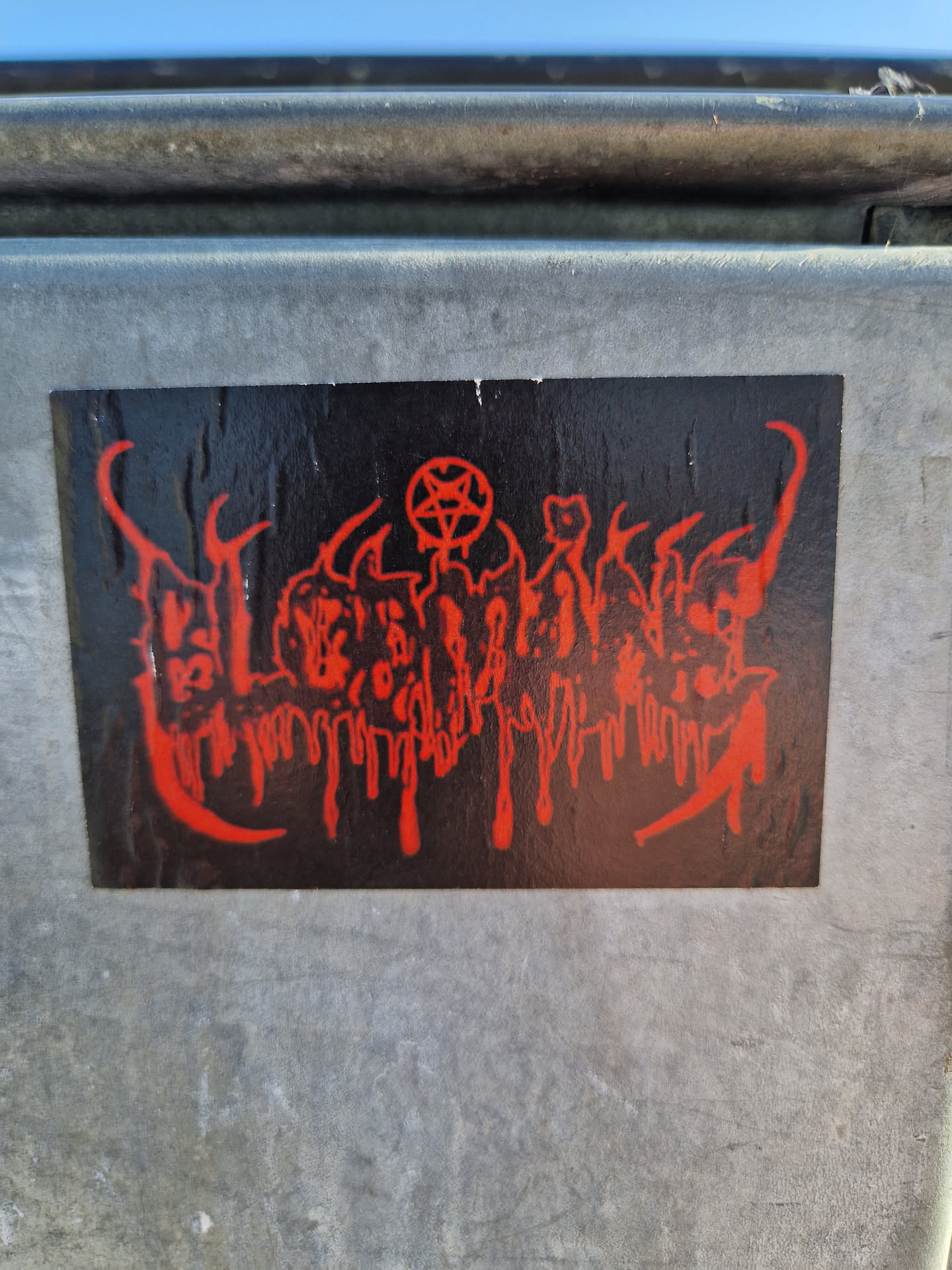 A black and red sticker with a design that appears to be inspired by occult or metal music imagery.  The sticker features dripping red elements and a pentagram. The text is illegible.