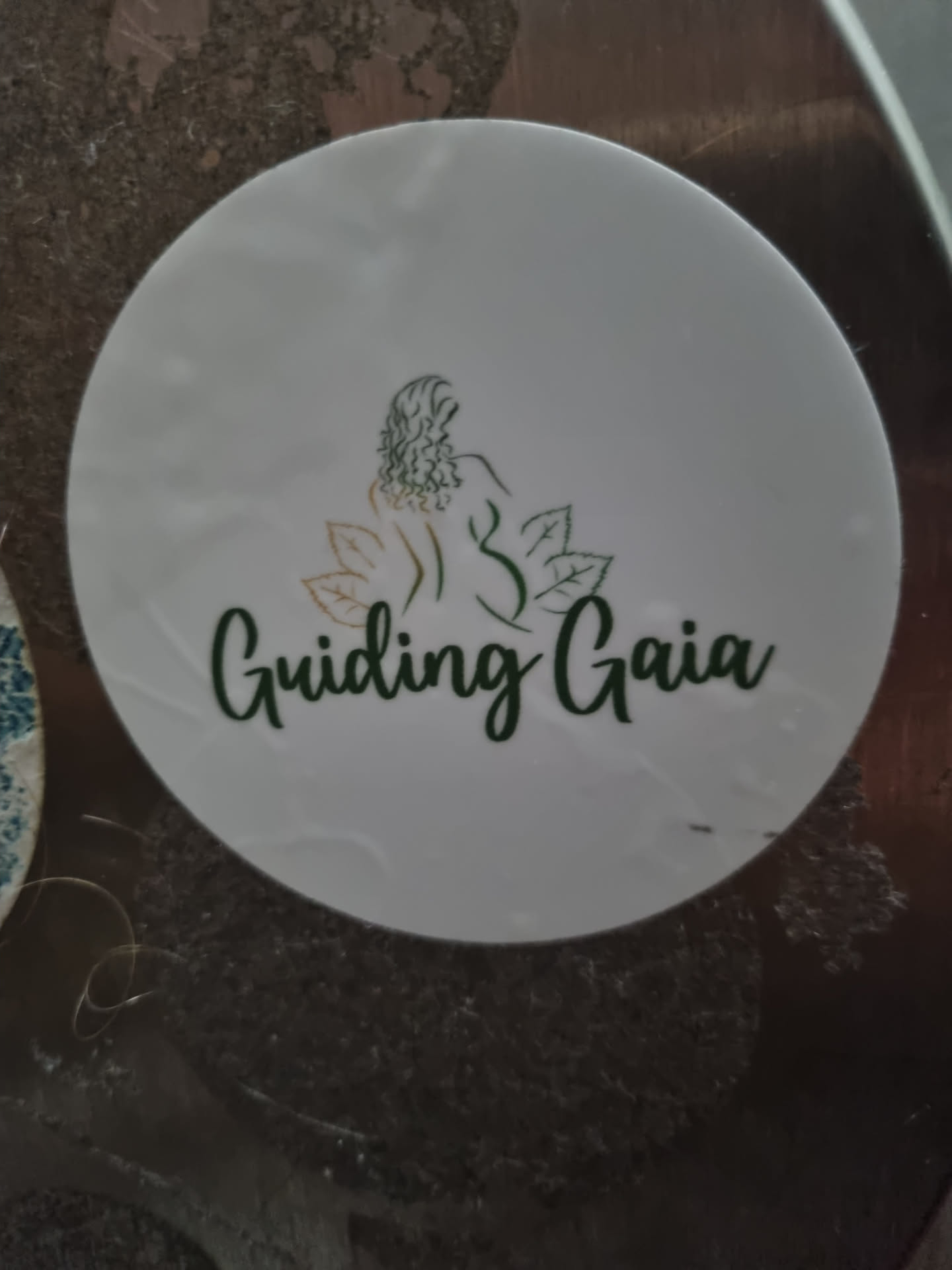 White round sticker with a logo of a woman sitting with leaves around her and the text Guiding Gaia written in a green cursive font.