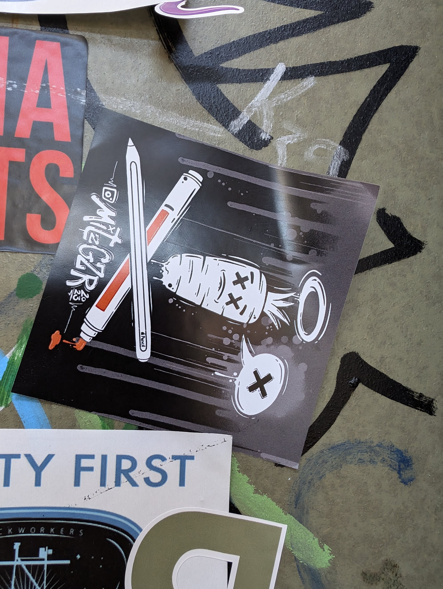 Black and white sticker with a design of two crossed markers and a dead carrot with an X on its face. The background is black with gray stripes. The artist's Instagram handle is @mitecar.