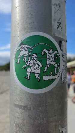 Street sticker A circular sticker on a green background with a white border. It depicts a humorous black-and-white comic illustration of three Swedish football players. In the center, a smiling player wearing a jersey labeled 'HIF' (Hammarby IF) charges forward dribbling a soccer ball. Flanking him in the background are two disoriented opponents labeled 'DIF' (Djurgårdens IF) and 'AIK', with motion lines depicting that they have been cleanly bypassed.