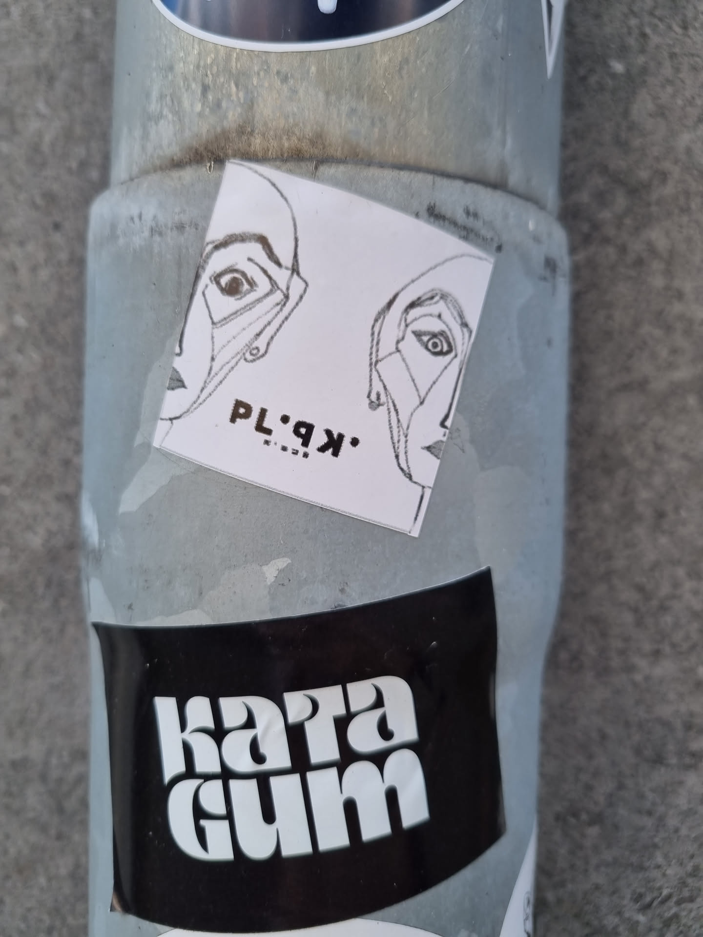 A small white square sticker with a black and white line drawing of two faces looking in opposite directions. The text 'PL·9K·' is visible below the drawing.  The sticker is attached to a metal pole, next to another sticker reading 'Kara Gum'.