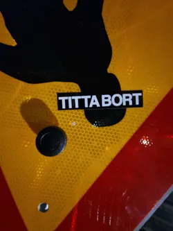 A black and white sticker with the text "Titta bort" (Swedish for "Look away") is affixed to a yellow and red traffic sign featuring a black silhouette of a person. The sticker is simple and minimalistic.
