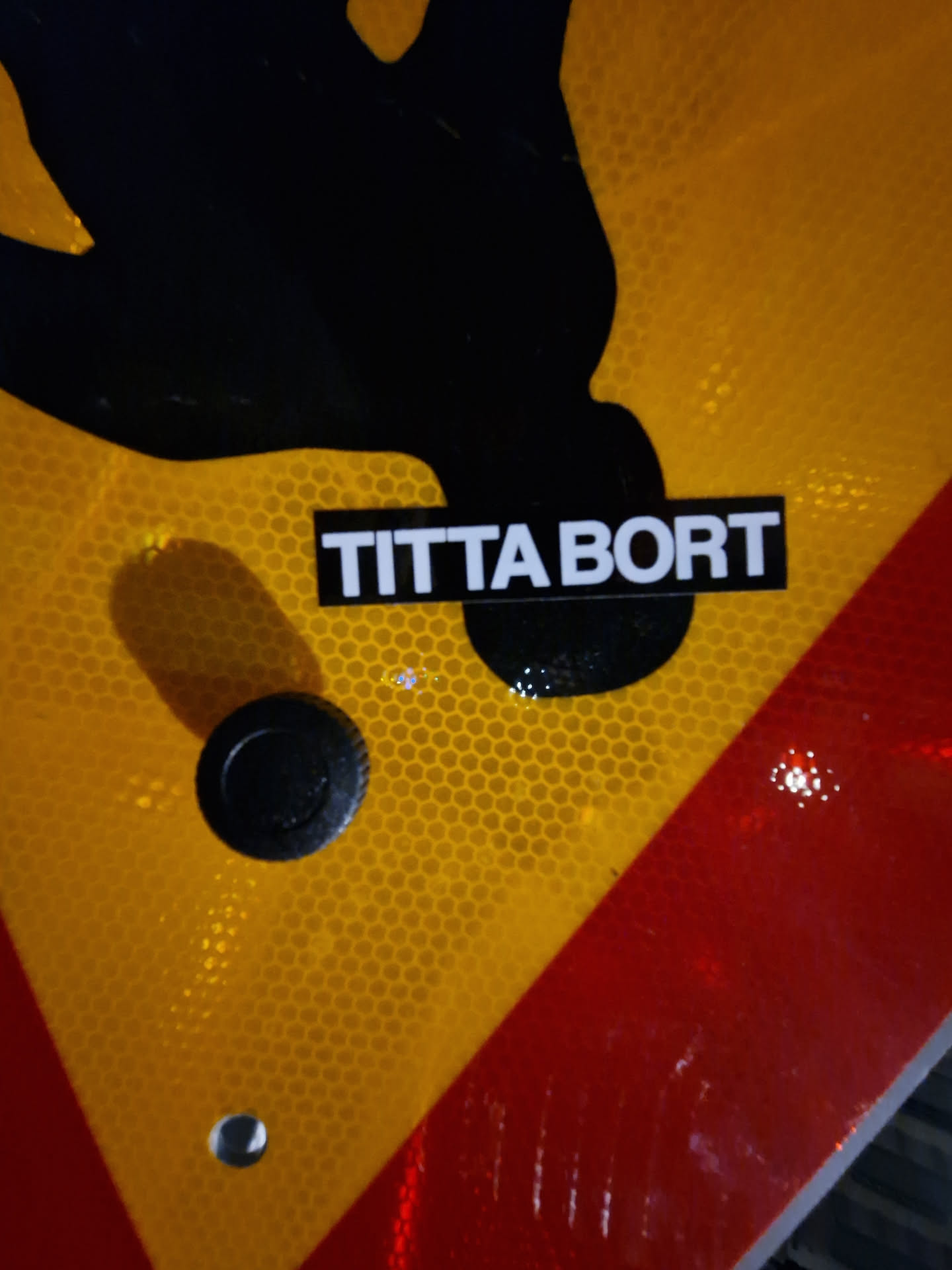 A black and white sticker with the text "Titta bort" (Swedish for "Look away") is affixed to a yellow and red traffic sign featuring a black silhouette of a person. The sticker is simple and minimalistic.