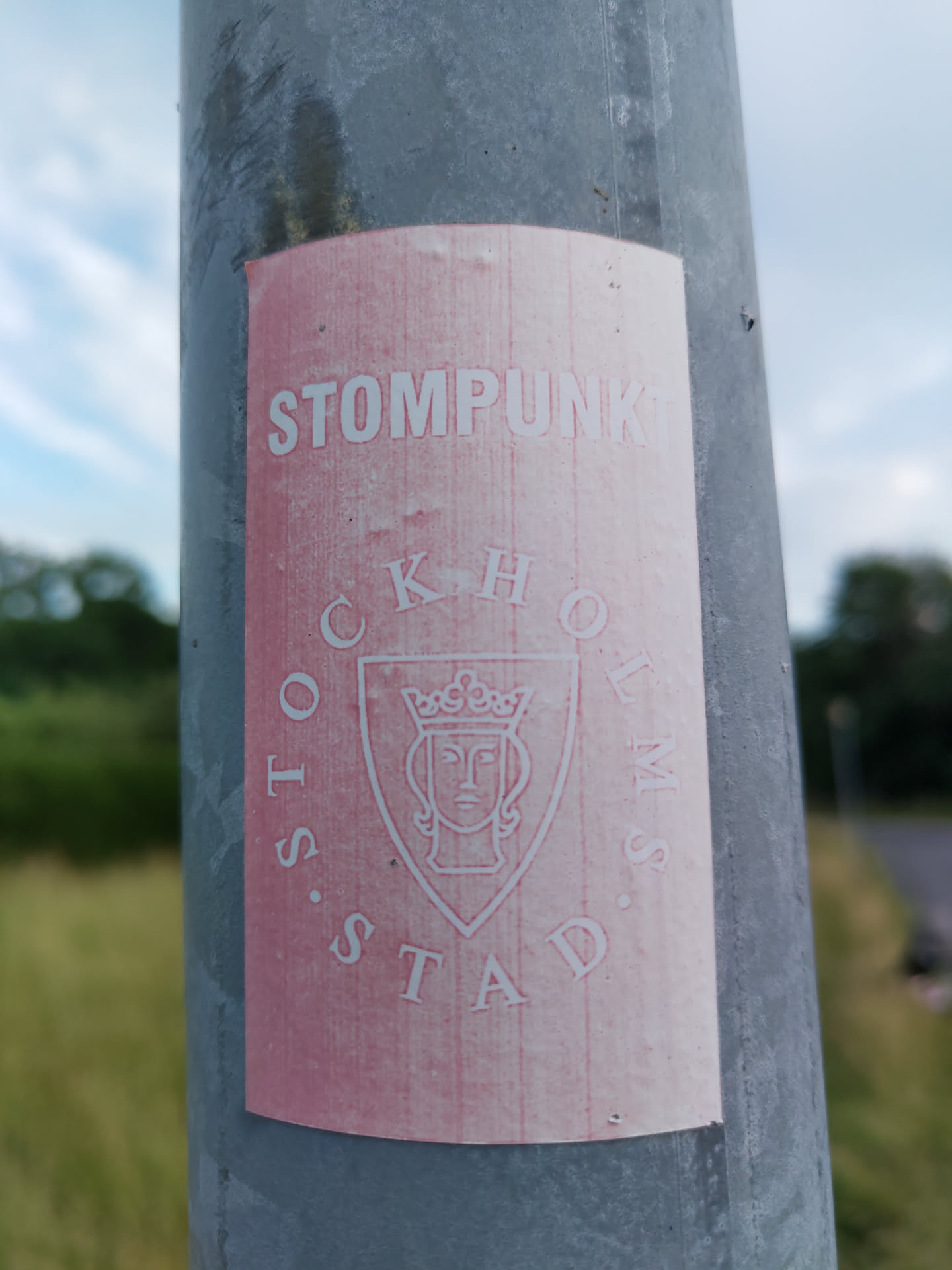 Pink and white sticker with the text "STOMPPUNK" at the top and a circular design with the text "STOCKHOLMS STAD" and a coat of arms in the center. The coat of arms shows a crowned woman's head. The sticker is attached to a gray pole.