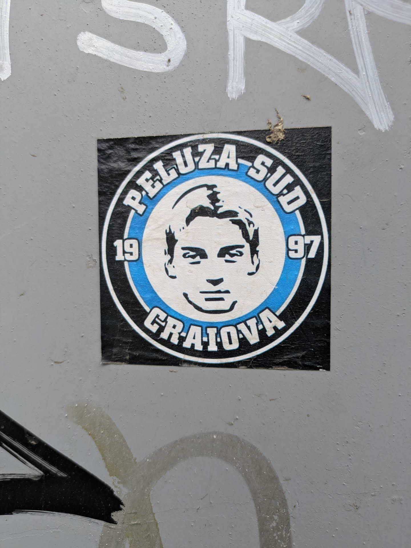 The sticker features a black and white portrait of a man inside a blue and white circle. The text 'PELUZA-SUD' and 'CRAIOVA' are printed in bold letters around the circle, with the years '19' and '97' flanking the portrait.