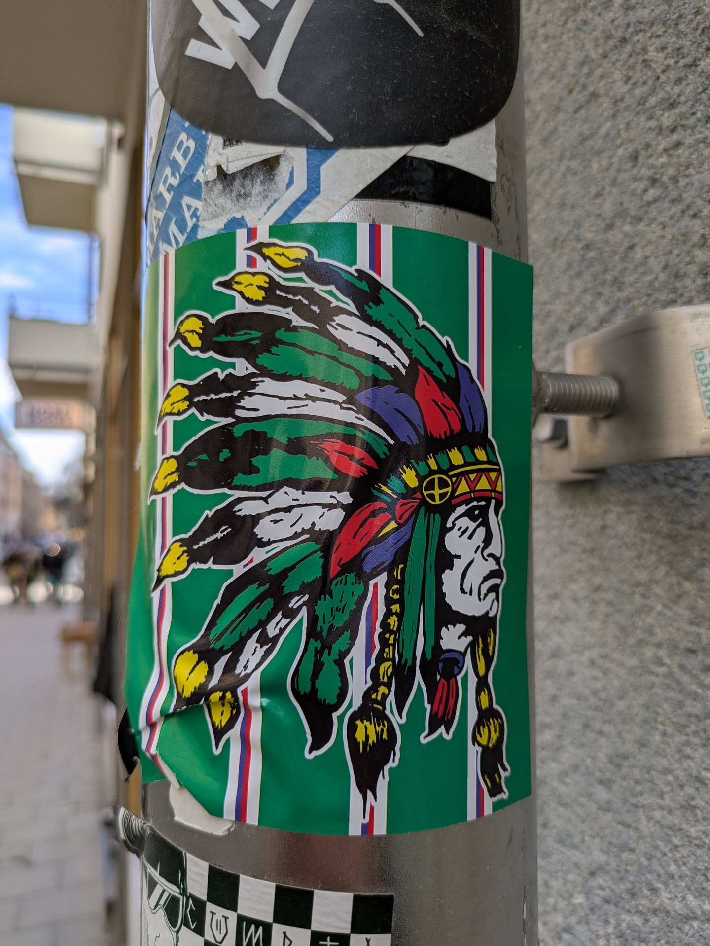 This sticker features a stylized portrait of a Native American chief wearing a colorful feathered headdress. The background has green, white, and red vertical stripes.