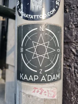 Black and white sticker with a hexagram shape in the center, surrounded by a compass rose. The text "KAAP A'DAM" is written below the design. The sticker is affixed to a pole.