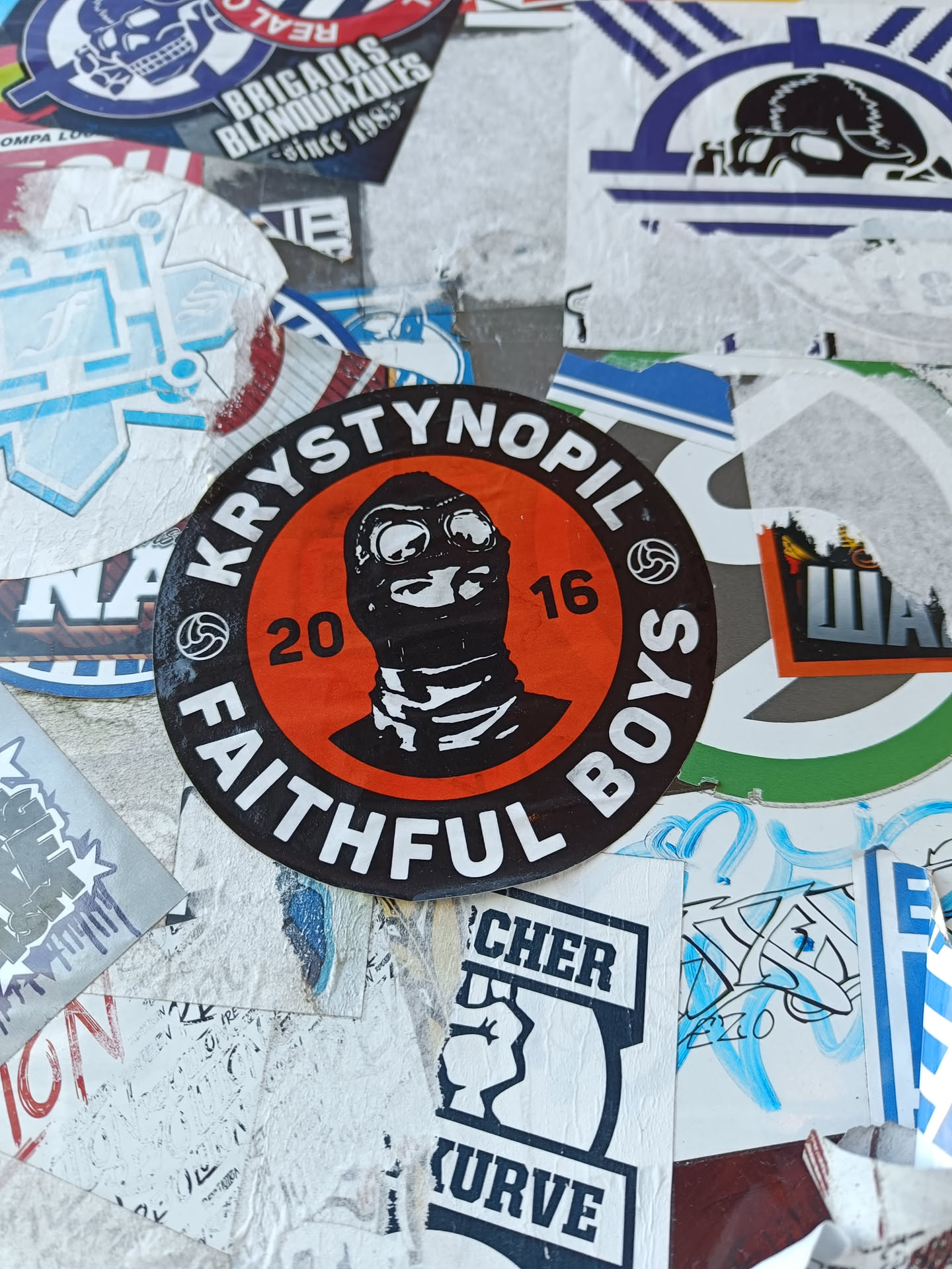 A round sticker with a black and orange color scheme. It has an illustration of a person wearing a balaclava and goggles in the center. The text "Krystynopil" and "Faithful Boys" are written around the illustration, with the numbers "20" and "16" flanking the illustration. The style is reminiscent of a logo or emblem.