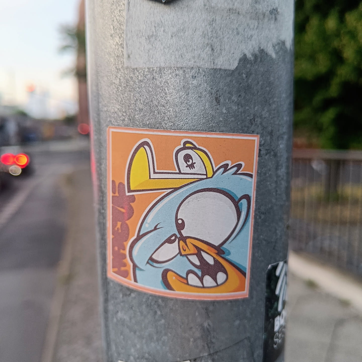 This sticker features a light blue bird with a large eye and a mischievous expression.  It's wearing a yellow hat with a small skull on it and is set against an orange background. The style is reminiscent of graffiti or street art.