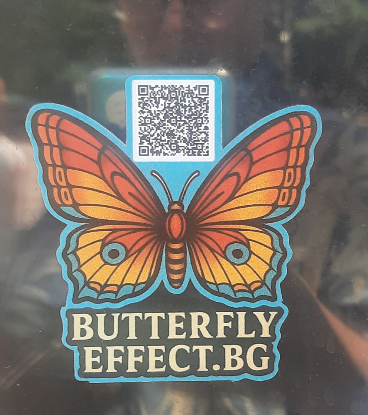 The central sticker features a stylized butterfly with wings in shades of orange, yellow, and reddish-brown, outlined by a distinct light blue border. The upper wings are predominantly orange with darker veins, transitioning to yellow and reddish-brown towards the lower parts. The lower wings are mainly yellow with orange and reddish-brown accents, including a small blue circle with a black dot on each. Above the butterfly's body, a black and white QR code is prominently displayed against a light blue background. Below the butterfly, the text "BUTTERFLY EFFECT.BG" is written in bold, light blue capital letters with a dark outline, matching the butterfly's border. The overall design has a clean, somewhat geometric aesthetic.