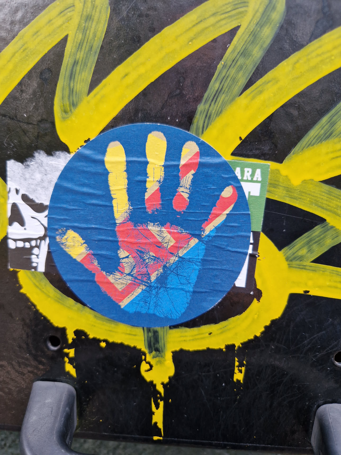 A blue circular sticker with a colorful handprint on it. The sticker is placed on a dark surface with yellow graffiti in the background. 