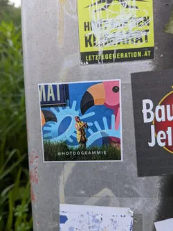 The sticker depicts a colorful scene where a person, dressed in a red-hooded hot dog costume, stands in a grassy foreground. Behind them is a brightly colored mural featuring a large pink flamingo head with a black beak on the right, and an abstract light blue pattern that resembles splashing water or stylized sun rays on a darker blue background. The person's hands are clasped together in front of them. The overall image has a playful and artistic feel.