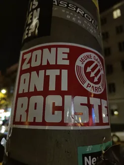 A red and white sticker with the text "ZONE ANTI RACISTE" and a logo that says "JEUNE GARDE PARIS". The sticker is affixed to a pole.