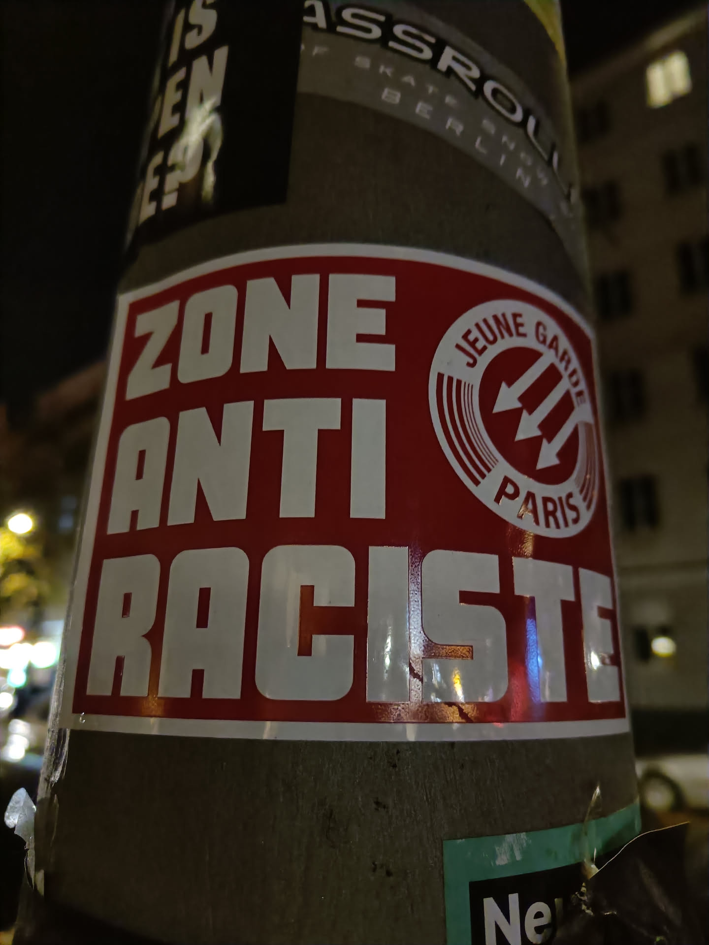 A red and white sticker with the text "ZONE ANTI RACISTE" and a logo that says "JEUNE GARDE PARIS". The sticker is affixed to a pole.