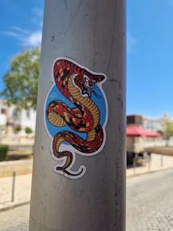 A sticker featuring a stylized illustration of a snake with tiger-like stripes, set against a blue circular background. The snake is coiled and appears aggressive. The sticker is affixed to a gray metal pole outdoors.