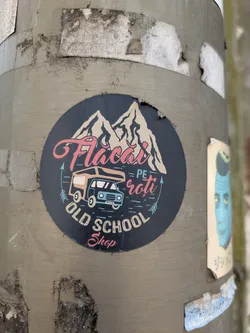 A circular sticker on a pole featuring a drawing of mountains, a camper van, and the text "Flacai pe roti Old school shop". The sticker is slightly torn on the upper side. The background color is dark grey, and the text is in pink and white.