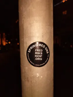 A black circular sticker on a metal pole.  The text on the sticker is in Swedish. It says "Spring med oss" (Spring with us), followed by "Fred Riks Hof .org", and a smaller line indicating "Sveriges Besta Langlöpare Sedan 1991" (Sweden's best long-distance runner since 1991).