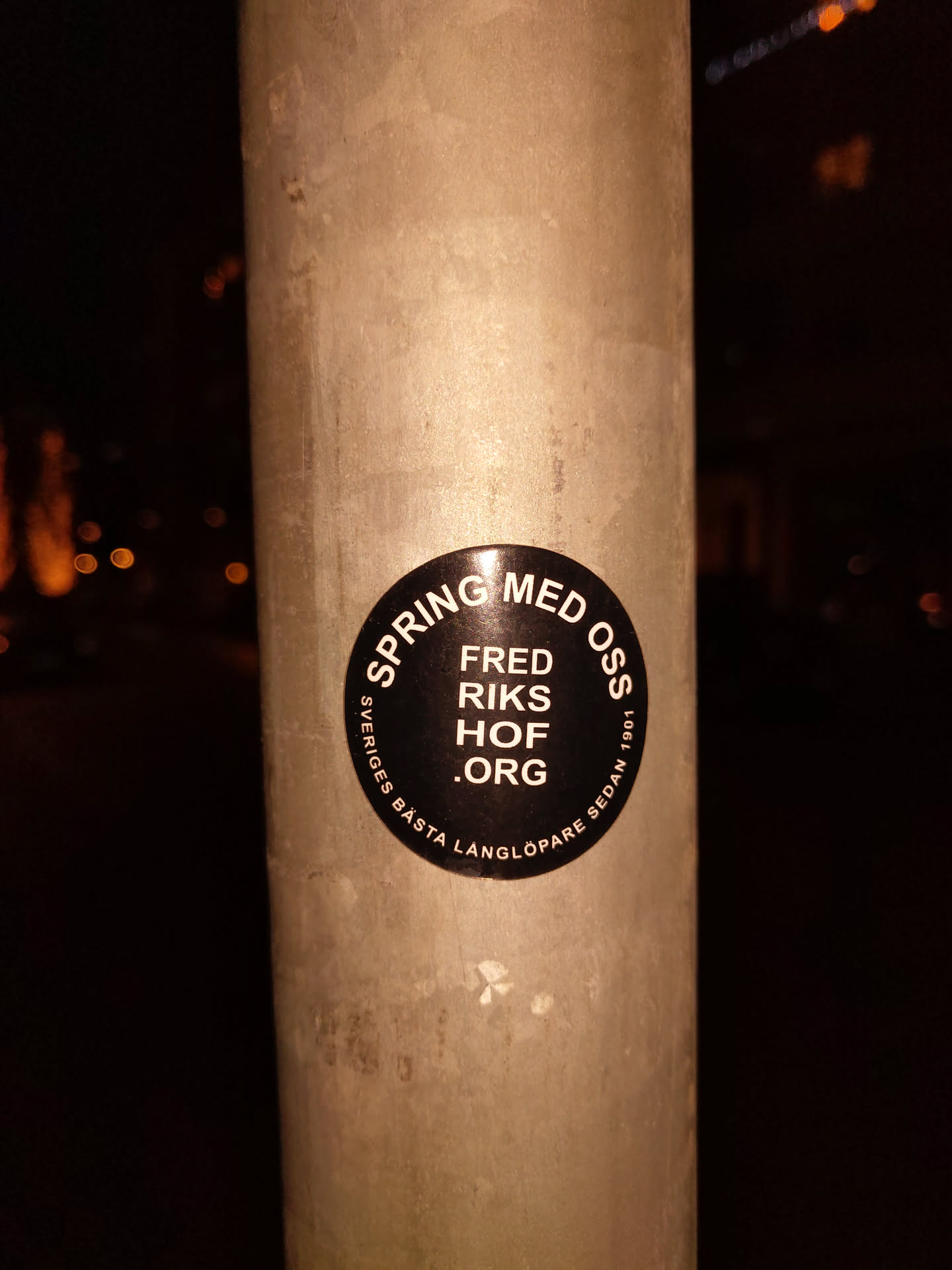 A black circular sticker on a metal pole.  The text on the sticker is in Swedish. It says "Spring med oss" (Spring with us), followed by "Fred Riks Hof .org", and a smaller line indicating "Sveriges Besta Langlöpare Sedan 1991" (Sweden's best long-distance runner since 1991).