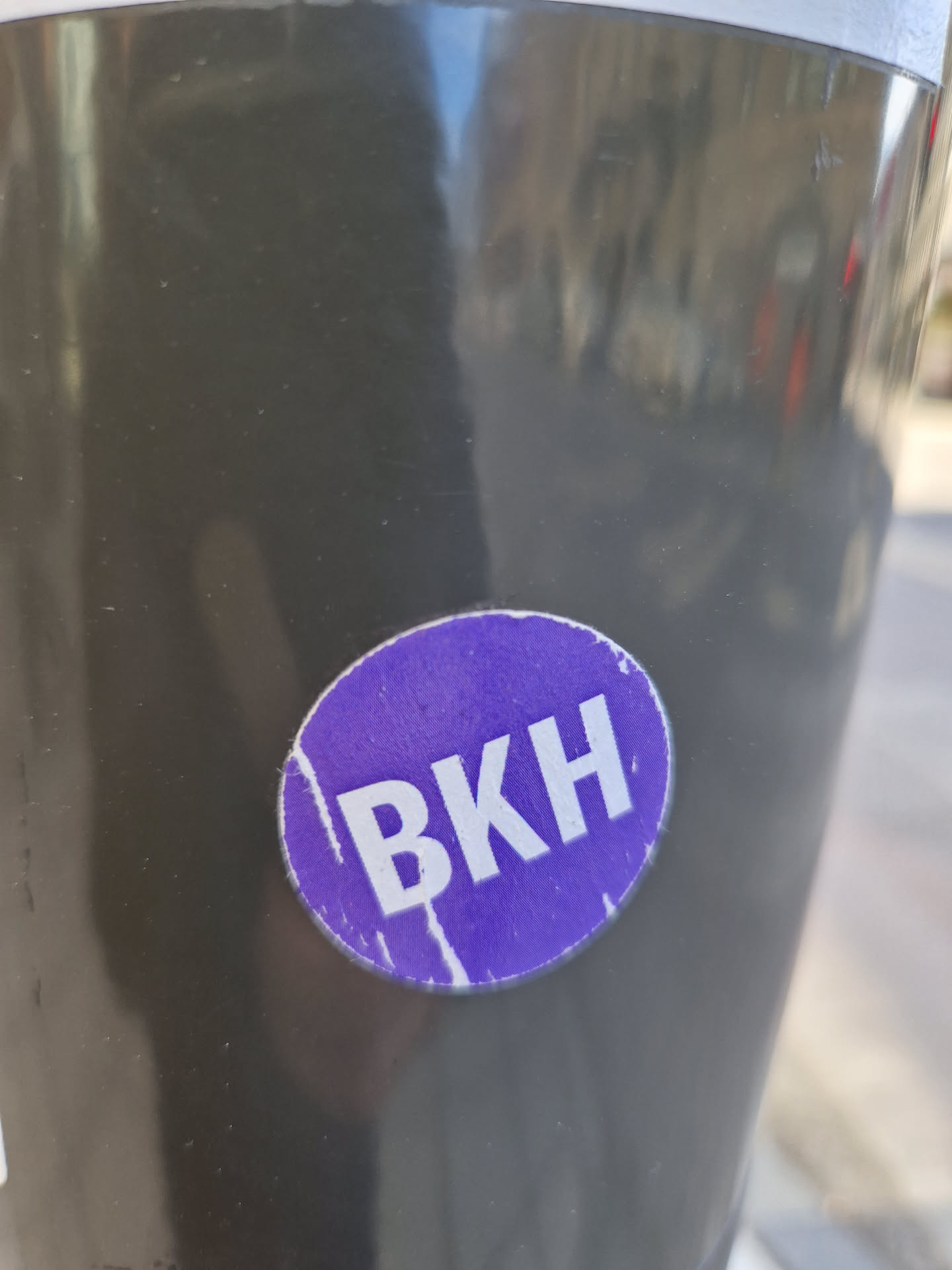 A round, purple sticker with the letters BKH in white capital letters. The sticker is slightly worn and has a few scratches on it. It's attached to a dark gray surface, possibly a pole or a piece of street furniture.