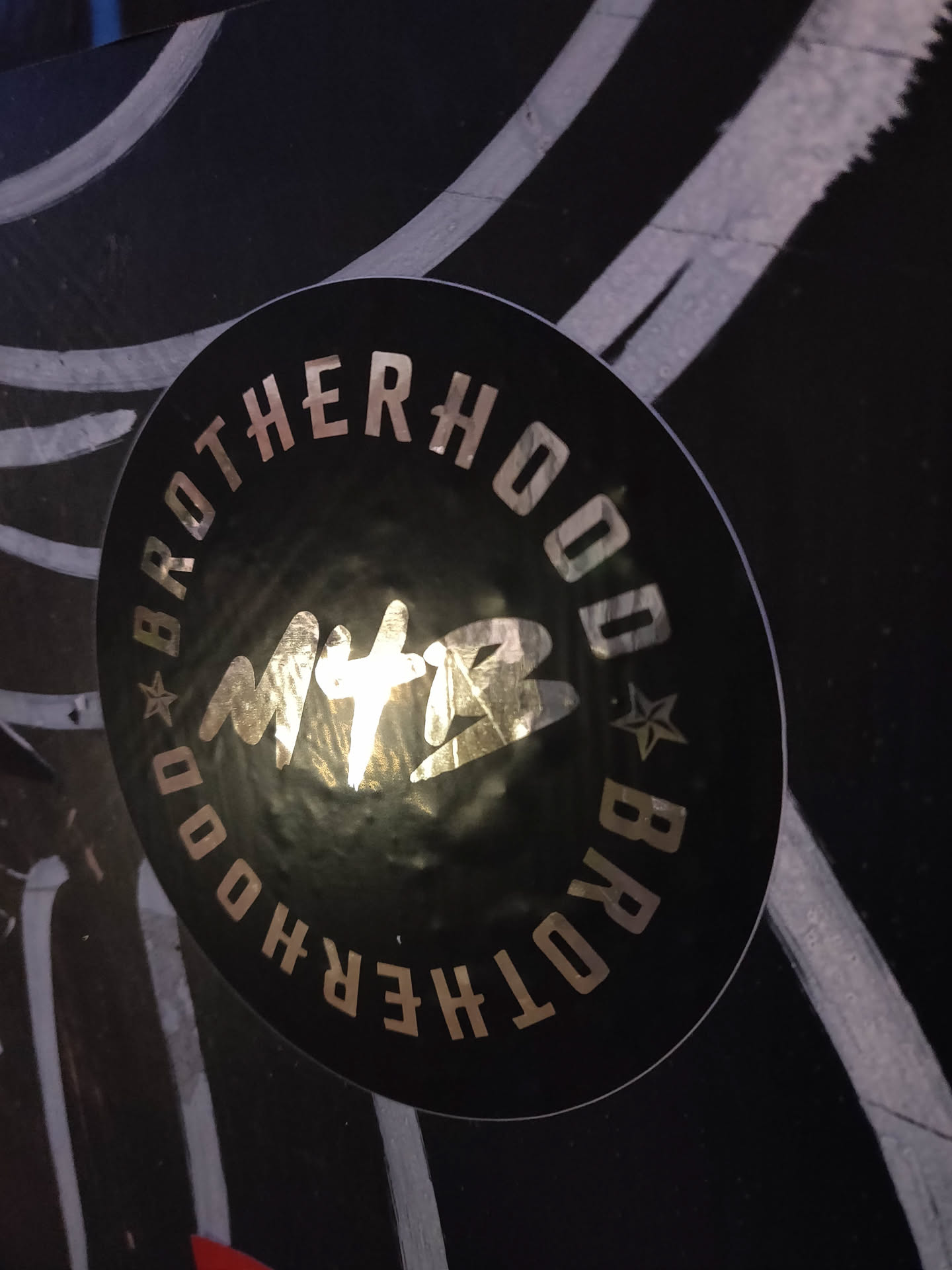 A black round sticker with the text "Brotherhood" written in a circular pattern around a central logo that appears to say "MTB". The text and logo are in a metallic gold color.