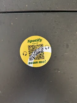 A round, yellow sticker features the Spotify logo along with the text "THT" and "scan me!". A QR code is prominently displayed, along with two headphone icons.