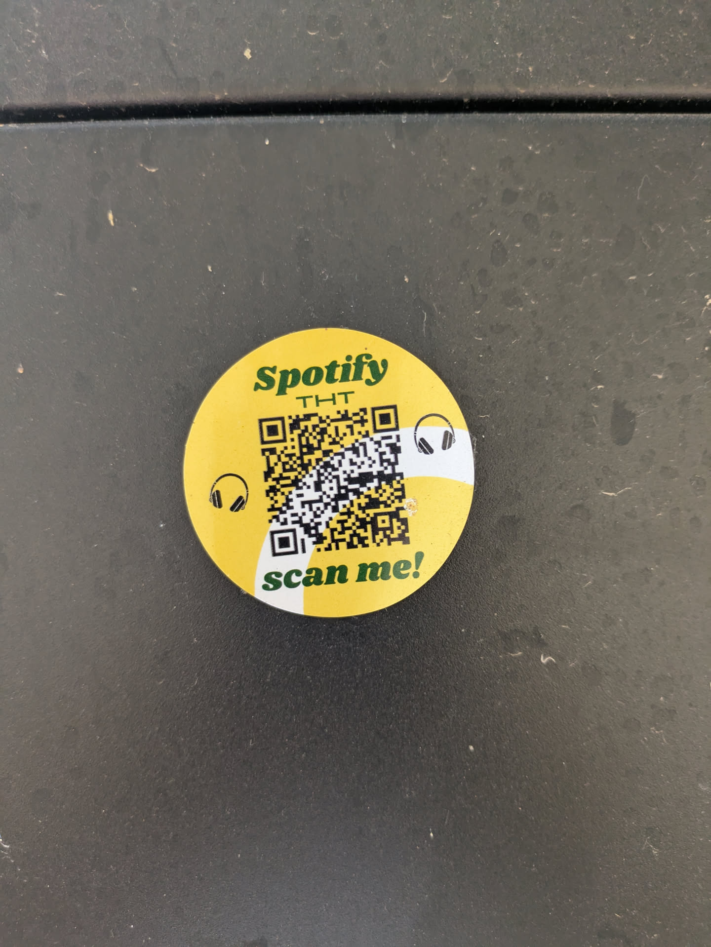 A round, yellow sticker features the Spotify logo along with the text "THT" and "scan me!". A QR code is prominently displayed, along with two headphone icons.